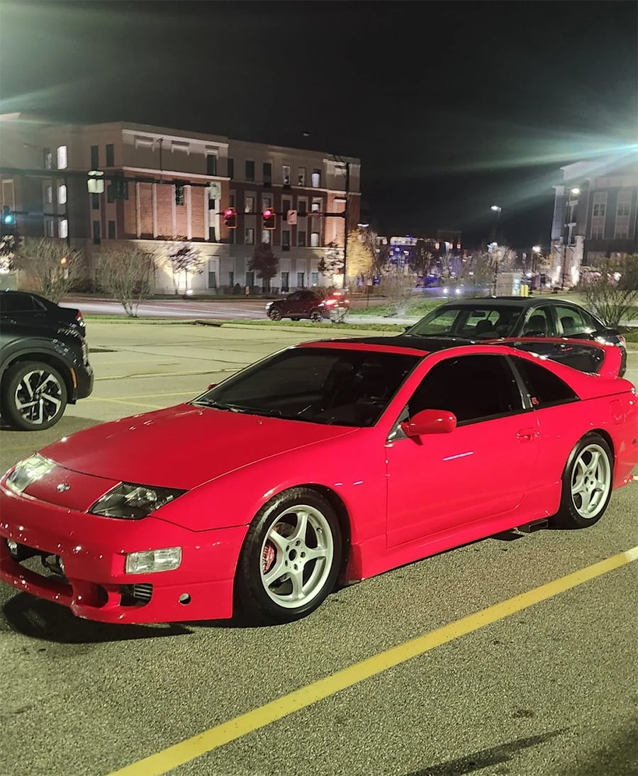 A bright red sports car is parked in a lot at night, with nearby buildings and streetlights illuminating the scene.