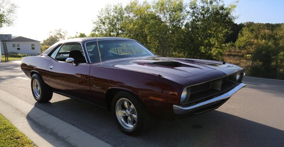 A classic dark purple muscle car with chrome wheels is parked on a sunlit suburban street near trees and a house.