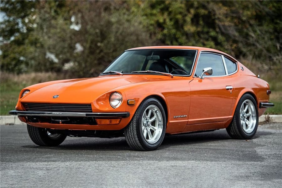 An orange classic sports car is parked on pavement, with trees and grass in the background. The car has a sleek, low profile and chrome wheels, with the driver’s side visible.