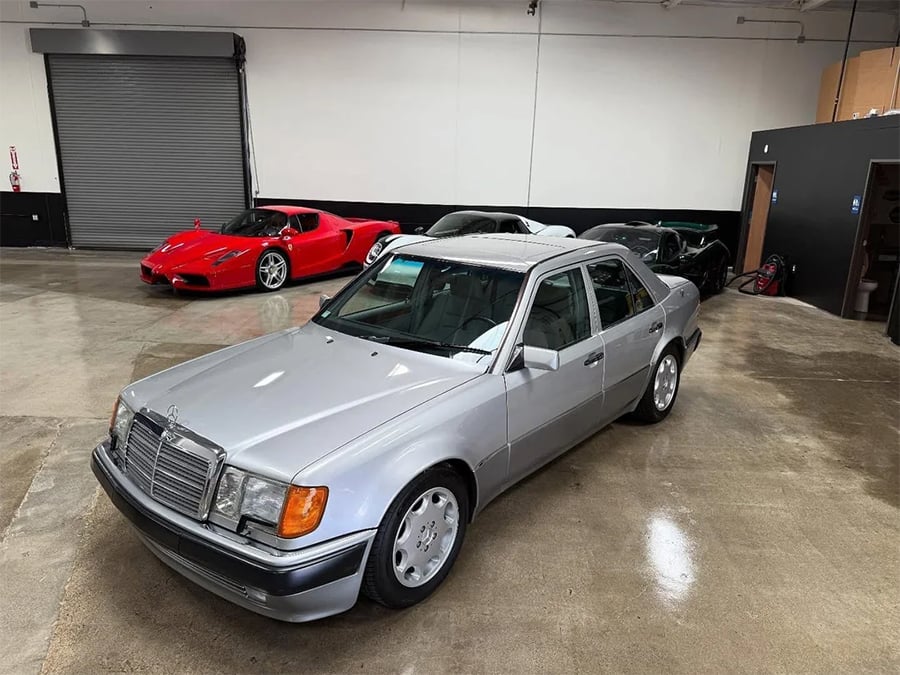A silver Mercedes-Benz sedan is parked indoors on a polished concrete floor. Behind it, a red sports car and two black cars are also parked. The space has white walls and a closed rolling garage door.