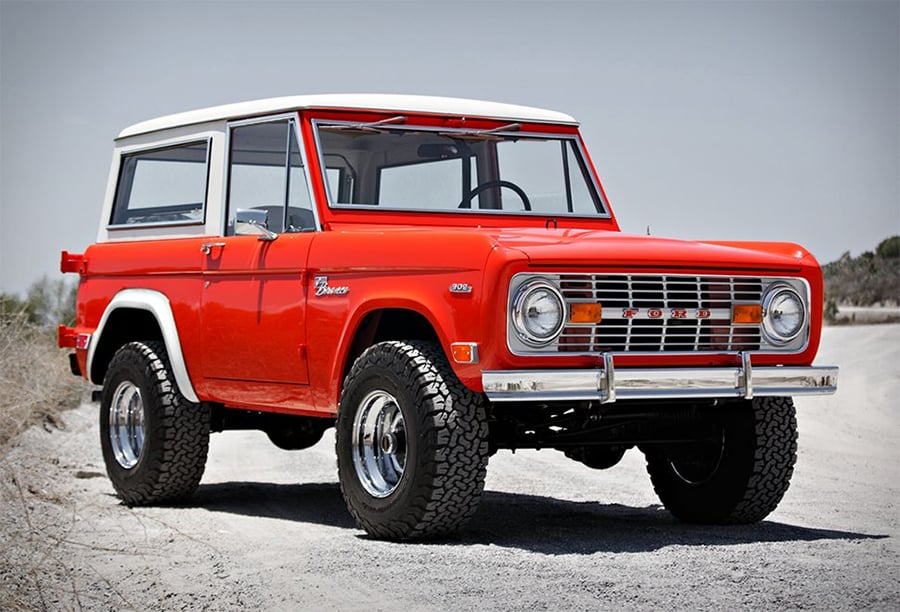 A classic red Ford Bronco SUV with a white roof is parked on a gravel road under a clear sky. The vehicle has large off-road tires and chrome accents, showcasing a vintage design.