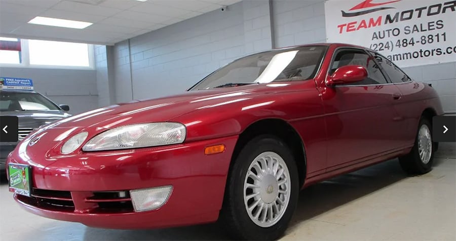A red Lexus SC coupe is parked indoors at an auto dealership. The car is facing left, with shiny paint and silver wheels visible, and dealership signs are on the wall in the background.
