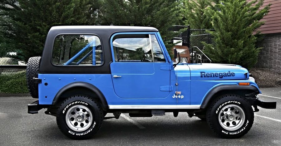 A blue Jeep Renegade with a black hardtop is parked in a lot, with trees and a fence in the background.