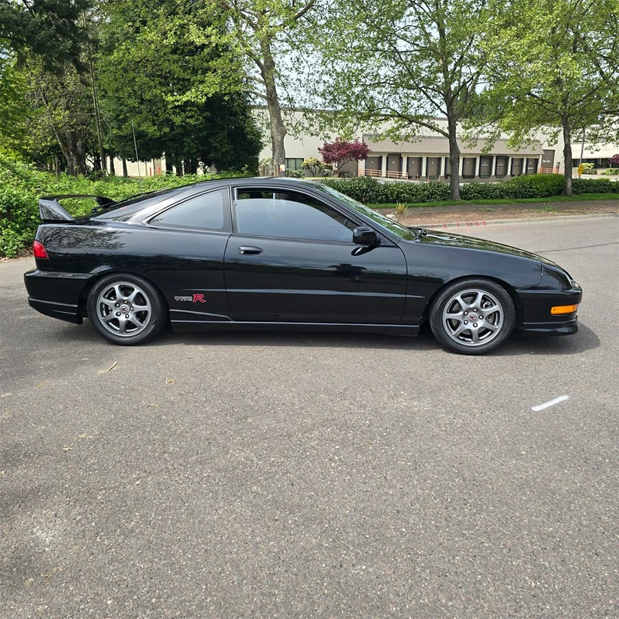 A black two-door sports car with a rear spoiler is parked on asphalt in a lot surrounded by trees and office buildings.