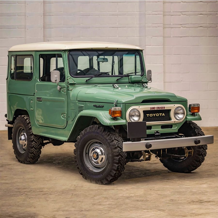 A vintage green Toyota Land Cruiser with a white roof is parked indoors on a concrete floor, in front of a white brick wall. The vehicle is clean and in excellent condition.