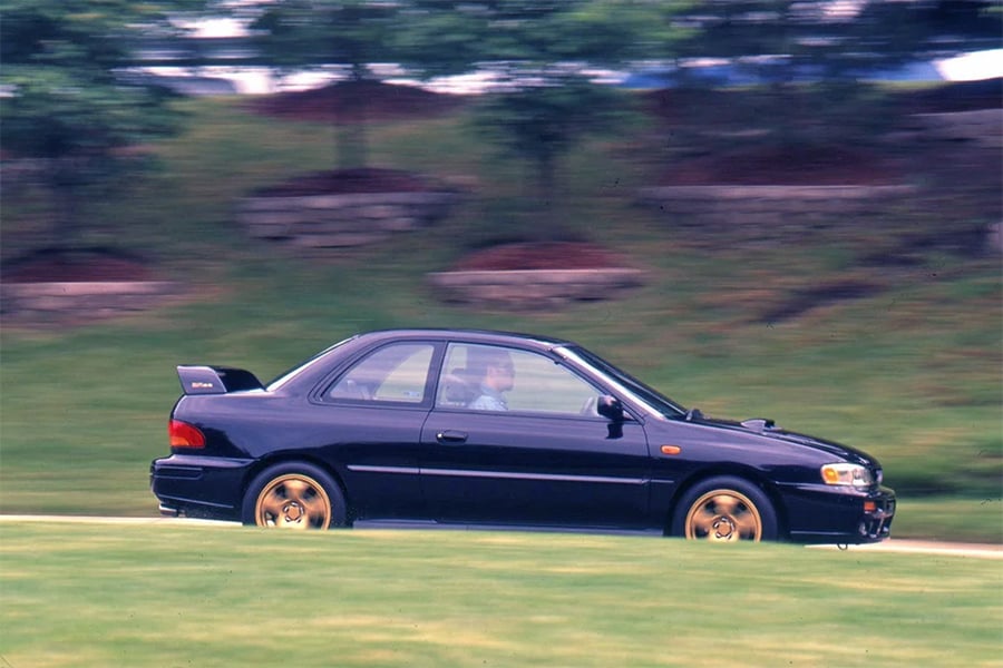 A dark blue sports coupe with gold wheels drives on a grassy road, with blurred green trees and stone walls in the background, suggesting motion.