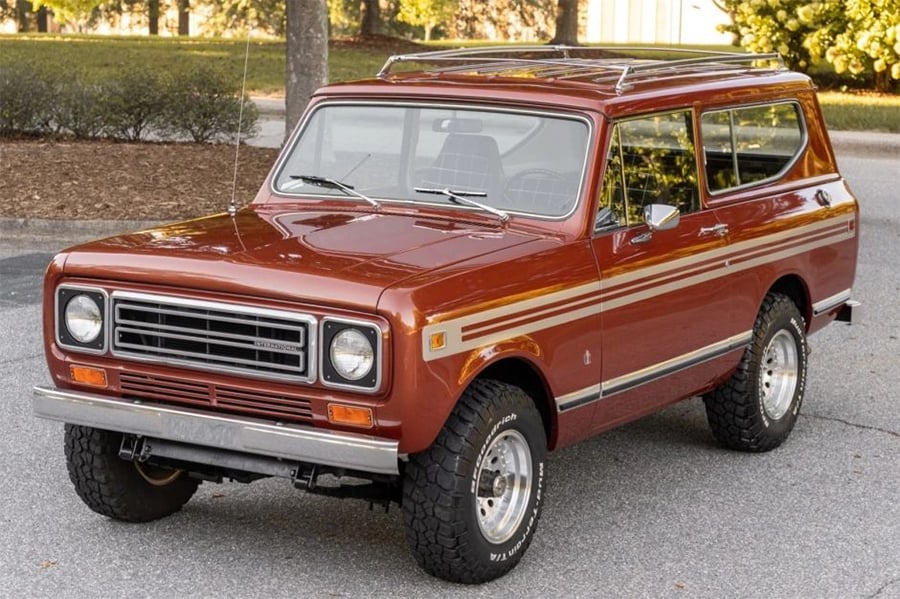 A vintage red SUV with white and yellow side stripes is parked on a street, featuring chrome bumpers, off-road tires, and a roof rack. Trees and bushes are visible in the background.