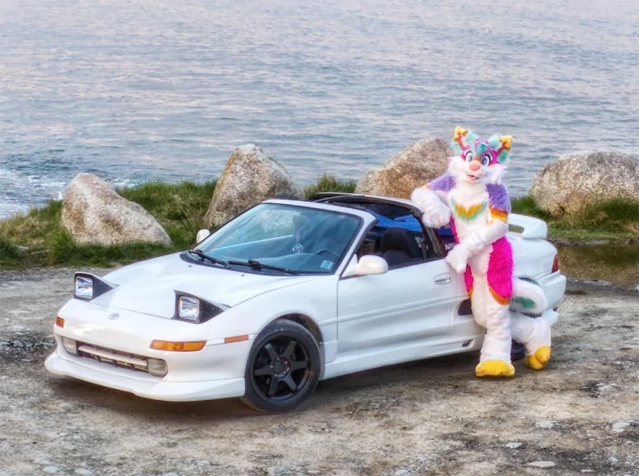 A person in a colorful fursuit stands next to a white sports car with pop-up headlights, parked by large rocks near a body of water.