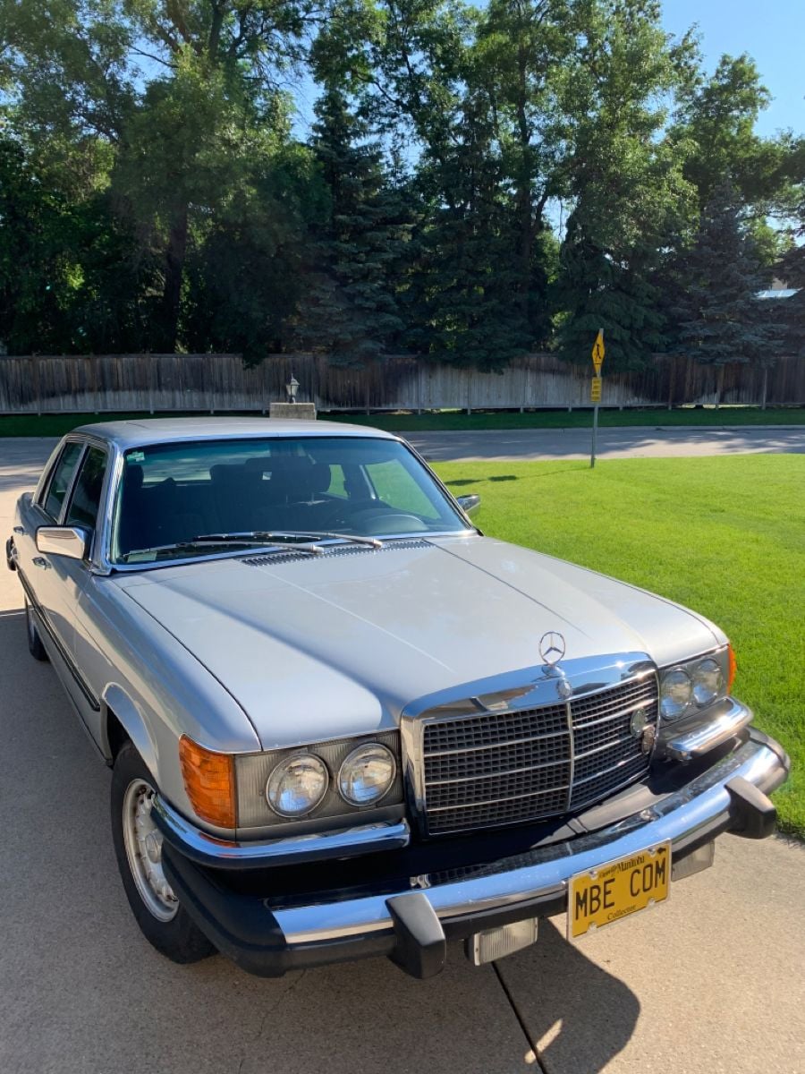 A silver classic Mercedes-Benz sedan is parked on a driveway in a suburban neighborhood, with a yellow license plate reading “MBE CDM.” Trees and a wooden fence are visible in the background.