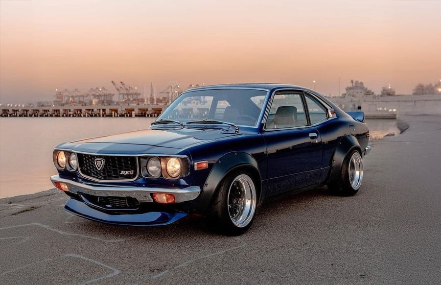 A classic dark blue Mazda RX-3 coupe is parked on a waterfront street at sunset, with industrial cranes and calm water in the background.