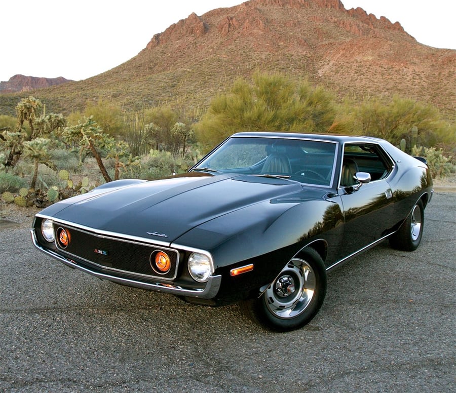 A classic black muscle car with round headlights is parked on a desert road with cacti and rocky hills in the background under a clear sky.