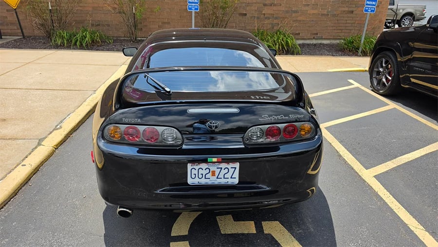 A black Toyota Supra is parked in a handicap parking spot, with its rear visible and a Missouri license plate reading “GGZ 1ZZ.” The car is next to a sidewalk and a brick building.