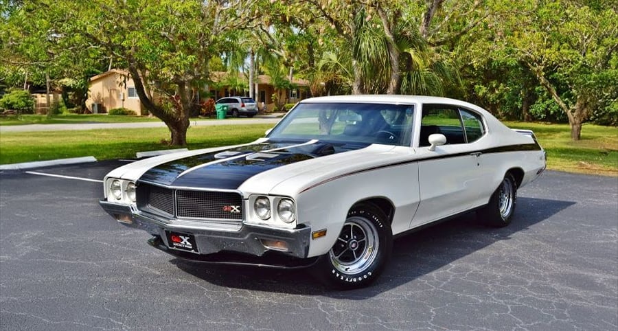 A classic white and black Buick GSX muscle car is parked on asphalt near trees and grass, with a house and other vehicles in the background.