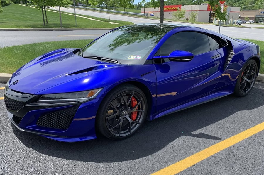 A blue sports car with tinted windows and black wheels is parked in a lot near grass and a shopping center on a sunny day.