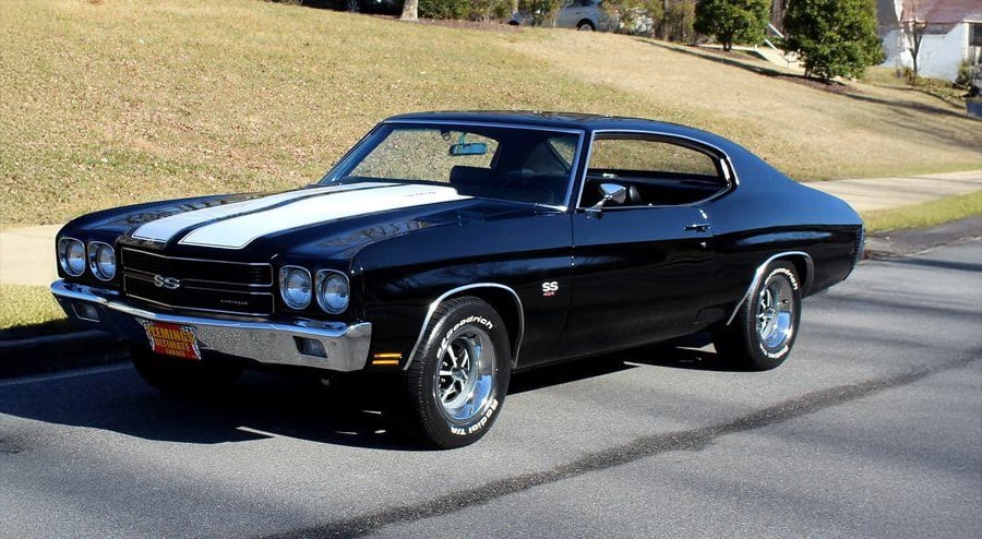 A black 1970 Chevrolet Chevelle SS muscle car with white racing stripes is parked on a suburban street, with grass and trees in the background.