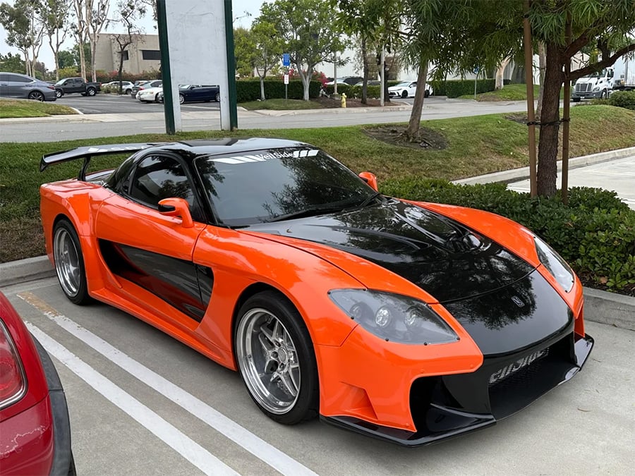 An orange and black sports car with a custom body kit is parked in a lot next to bushes and other cars, with buildings and trees in the background.