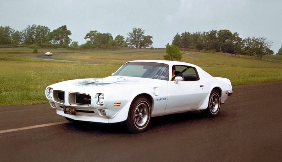 A white classic Pontiac Trans Am car with a blue firebird decal on the hood is parked on a paved road in a grassy countryside setting with trees in the background.