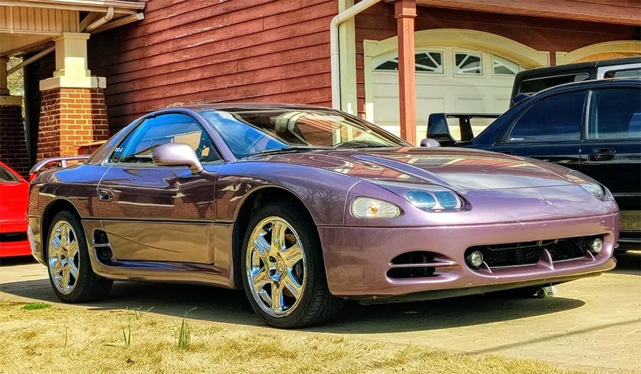 A purple sports car with chrome wheels is parked in a driveway in front of a house with wood siding and brick accents. Another black car is parked nearby.