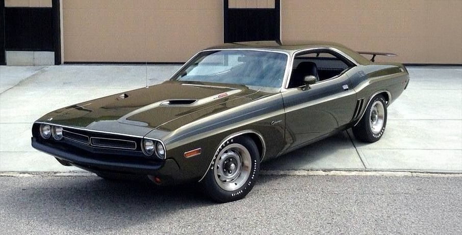 A classic dark green muscle car, likely a 1970s Dodge Challenger, parked on a driveway in front of tan garage doors.