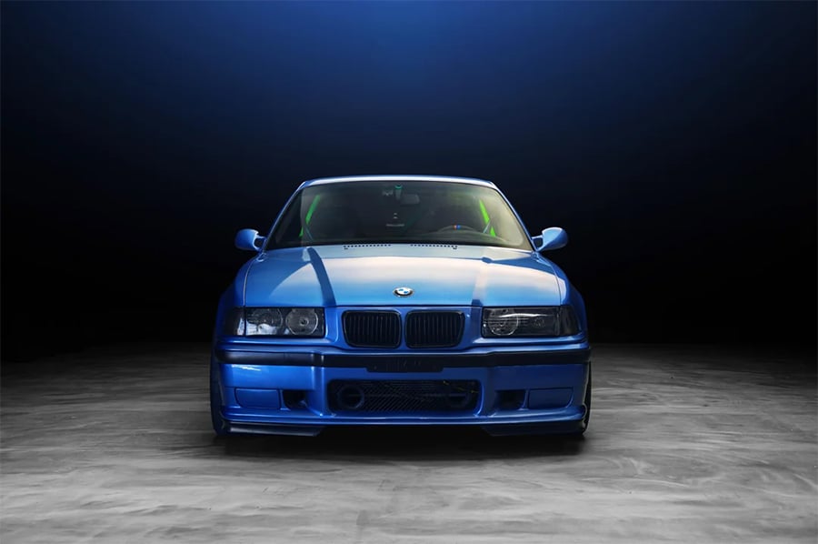 A blue BMW sports car is photographed head-on in a dark studio with a concrete floor and a blue gradient background. The headlights are off, and the car’s body reflects the studio lights above.