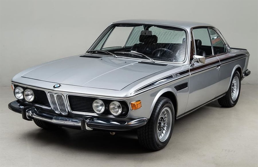 A silver classic BMW coupe with black detailing, chrome bumpers, and round headlights is parked in a studio with a plain background.