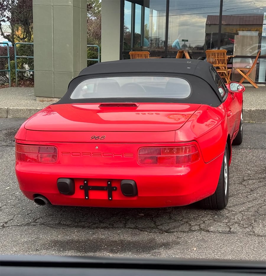 A red Porsche 968 convertible with a black soft top is parked outside a building with glass windows and outdoor wooden tables and chairs.