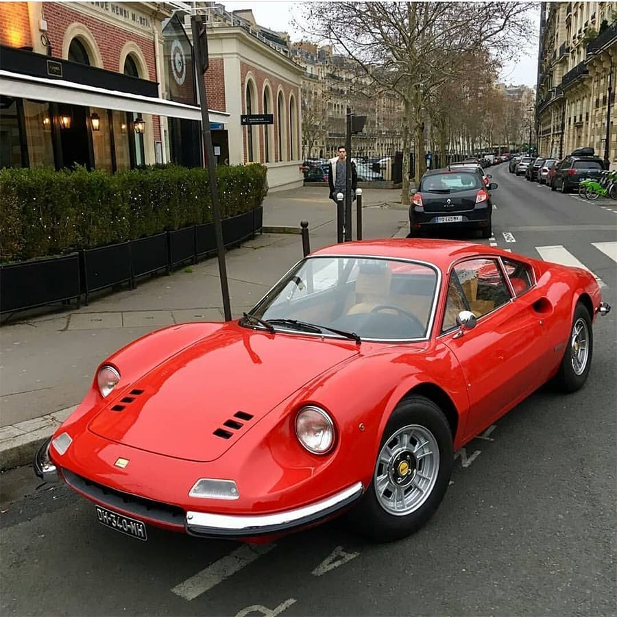 A classic red sports car is parked on a city street lined with historic buildings, with trees and a café visible in the background.