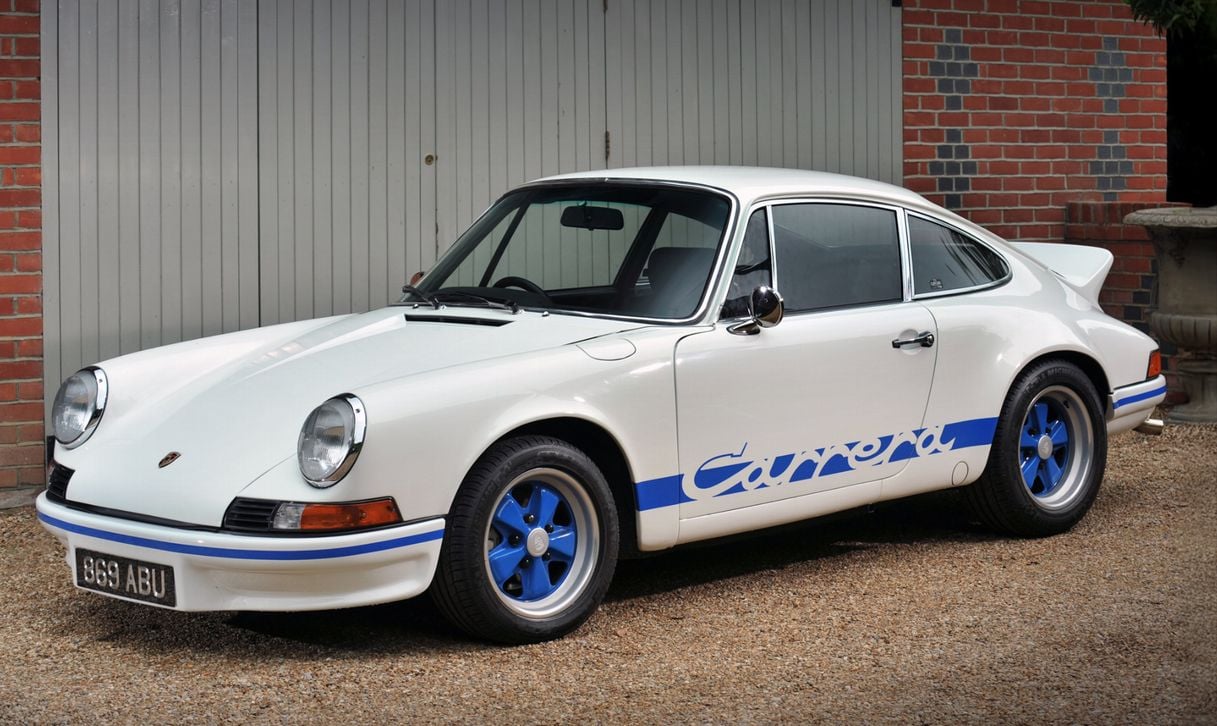 A classic white Porsche 911 Carrera with blue accents and blue wheels is parked on gravel in front of a brick building and a gray garage door.