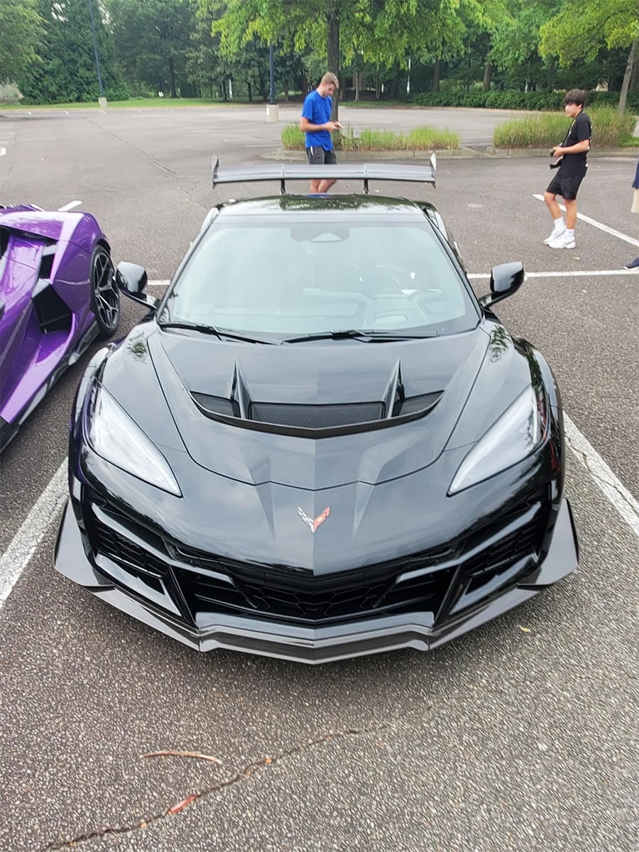 A black Chevrolet Corvette sports car with an aggressive front design is parked in a lot next to a purple car. Three people are standing nearby, and there are trees in the background.