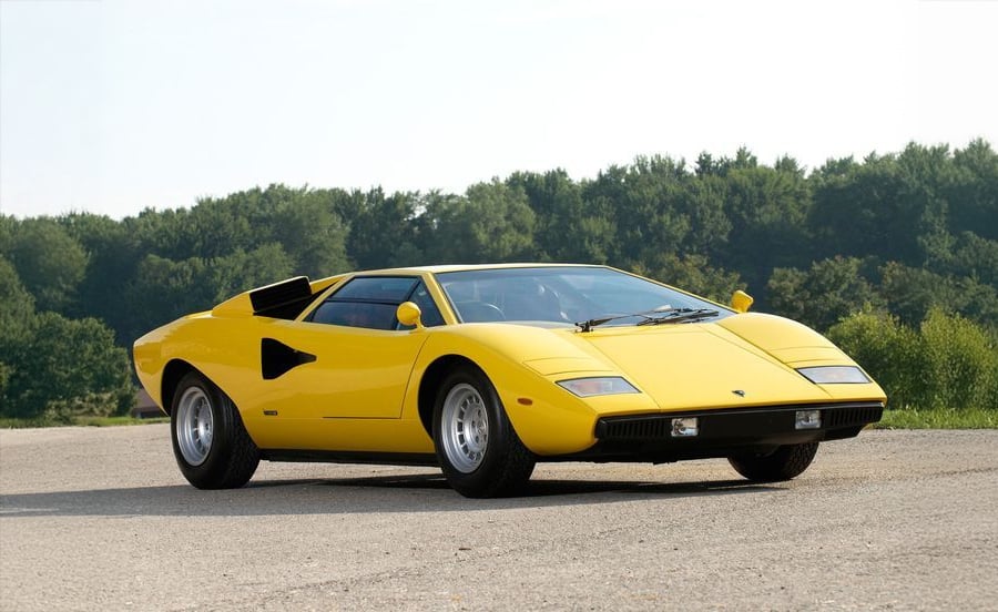 A yellow classic sports car with a low, angular design is parked on a paved surface, with green trees and a clear sky in the background.