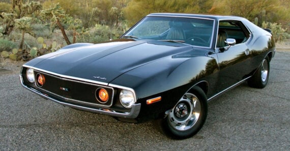 A classic black muscle car parked on a road, with desert plants and cacti in the background. The car has a sleek design, round headlights, and shiny chrome accents.