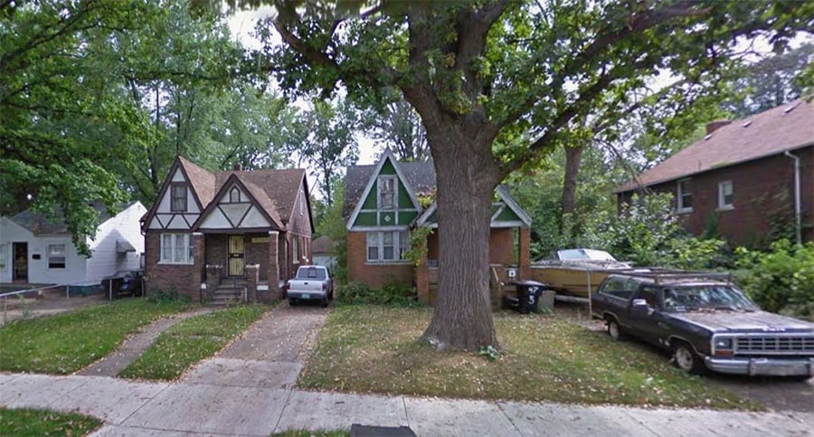 Two small Tudor-style houses with steep roofs stand side by side under a large tree. Several cars are parked in front, and greenery surrounds the homes on a quiet residential street.