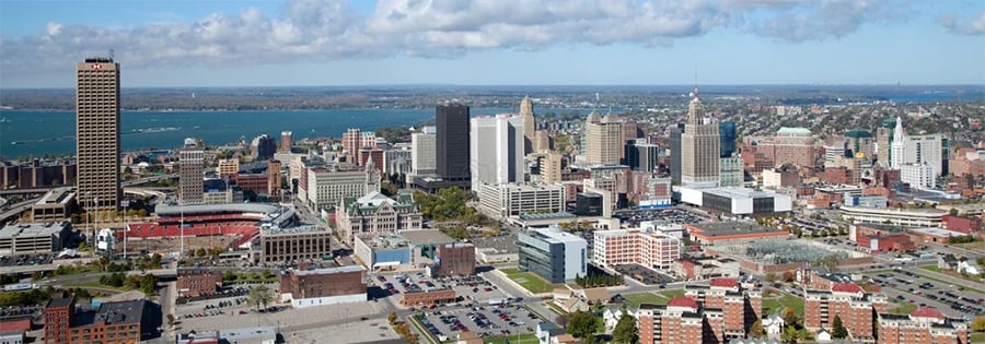 A panoramic view of a city skyline with tall buildings, a river or lake in the background, and partly cloudy skies. The foreground includes streets, parking lots, and low-rise structures.