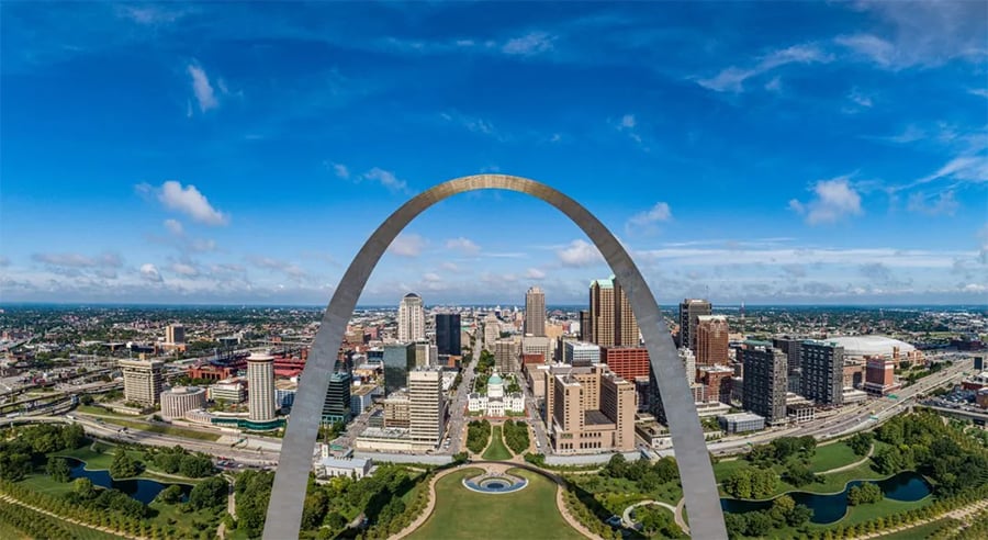 Aerial view of downtown St. Louis, Missouri, featuring the Gateway Arch in the foreground, with skyscrapers, green spaces, and a blue sky with scattered clouds.