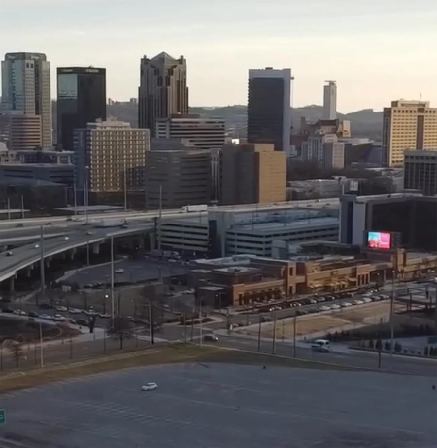 A city skyline with tall buildings, highways, a large parking lot in the foreground, and a bright digital billboard near street-level shops.