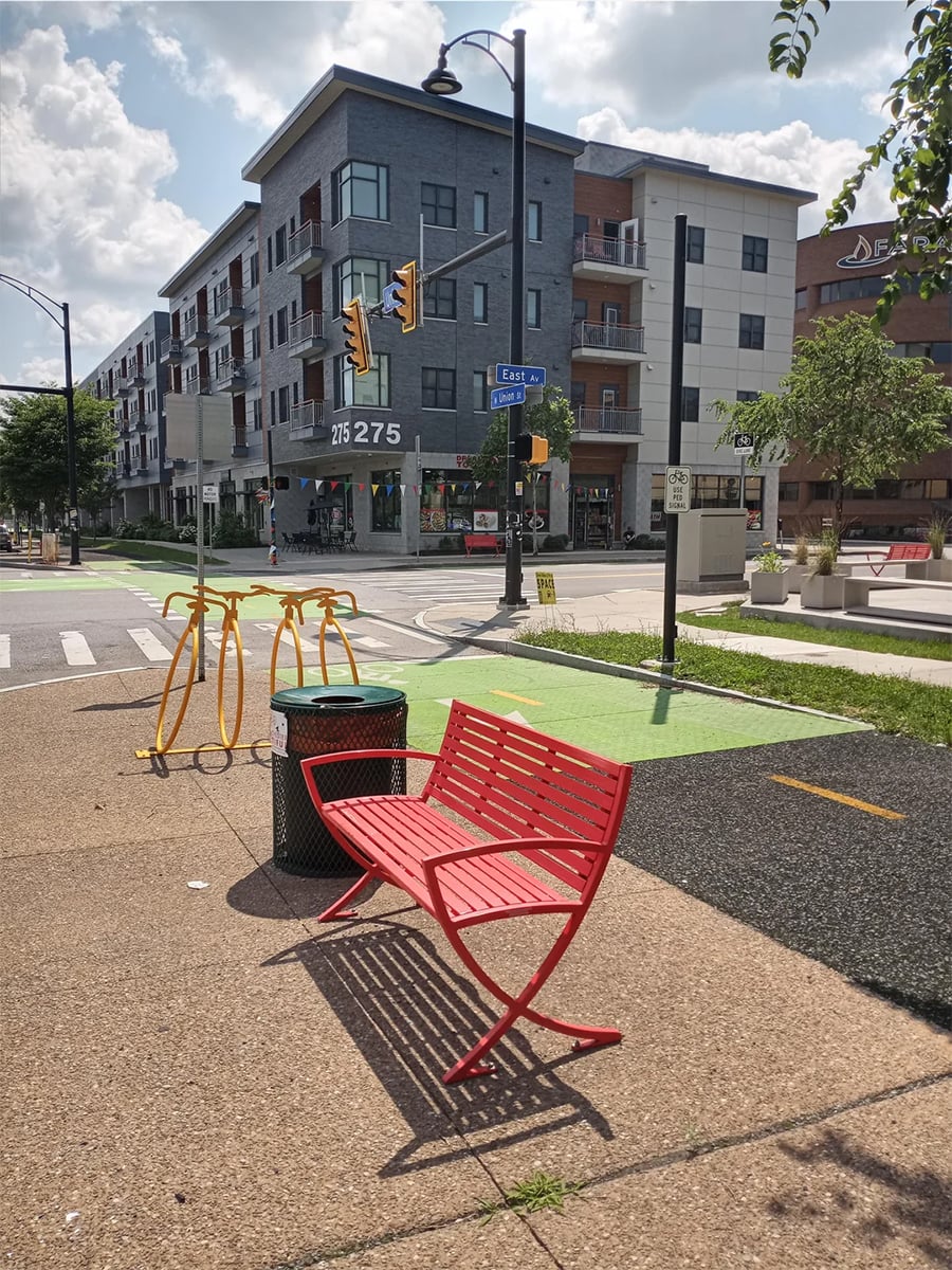 A bright red bench and bike rack sit on a sidewalk near a busy intersection, with a modern apartment building and green bike lane in the background on a sunny day.