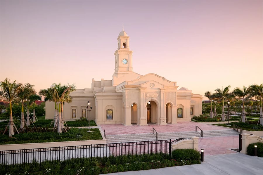 A tan, neoclassical building with a clock tower and arched entrance is surrounded by palm trees and landscaped gardens under a soft pink and purple sky at sunset.