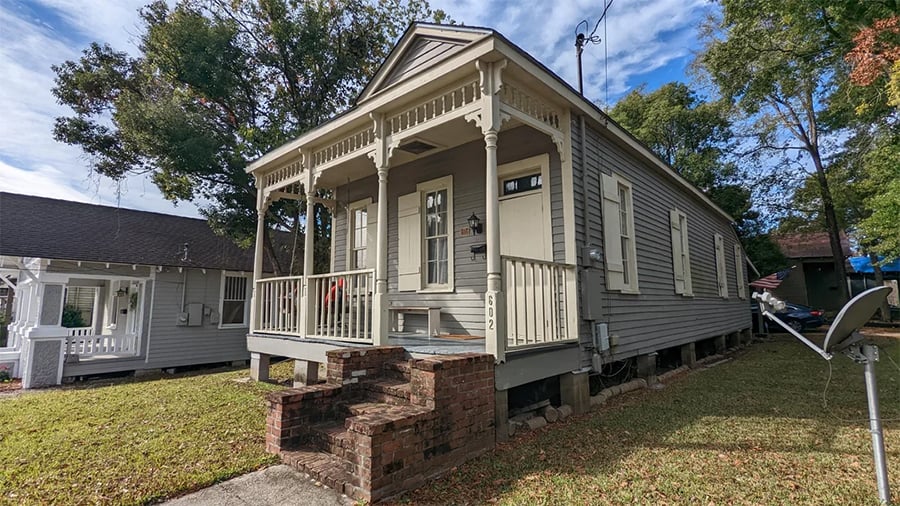 A small gray wooden house with a front porch, decorative trim, and brick steps. The yard is grassy, and a satellite dish is visible to the right. Trees and another house are in the background.