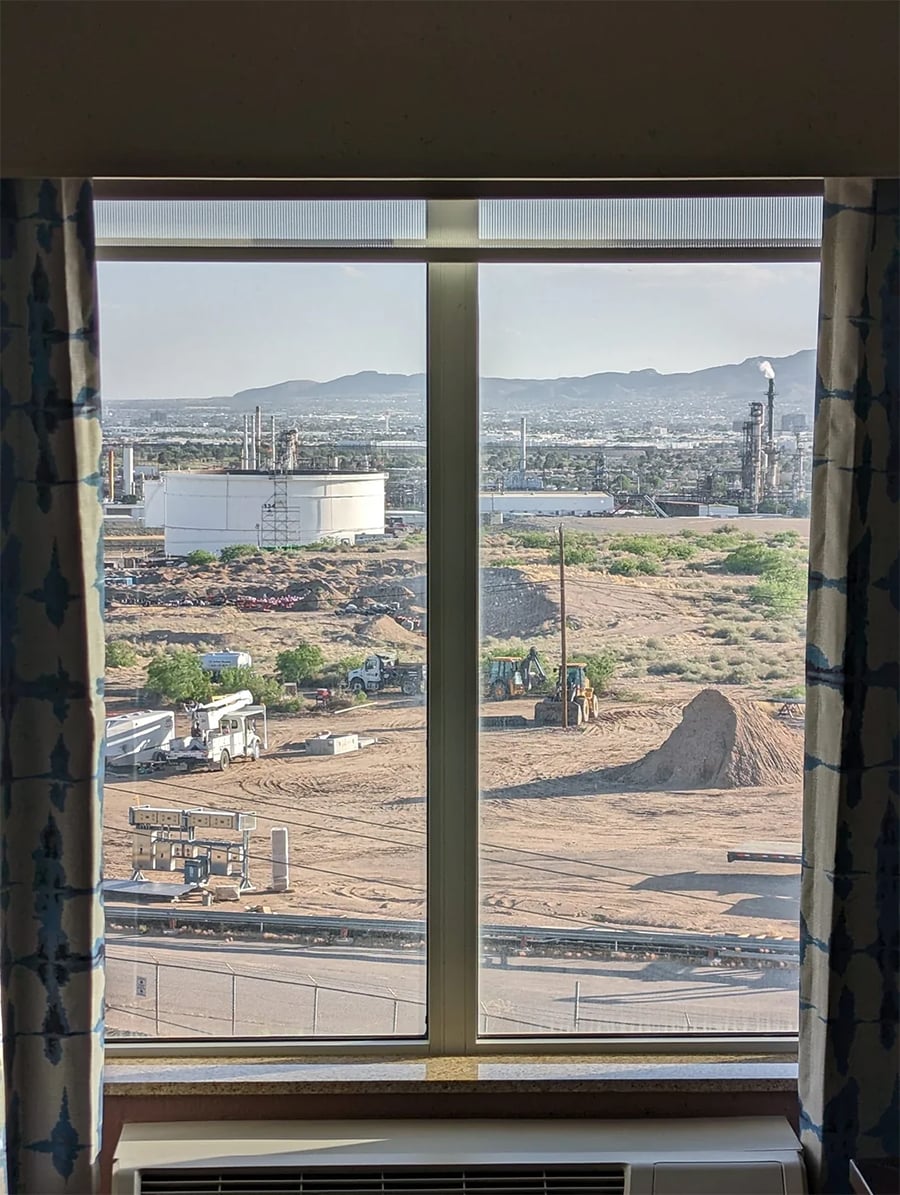 View through a window showing an industrial site with machinery, storage tanks, and pipes. Dirt ground, a small mound, mountains, and hazy cityscape are visible in the background. Curtains frame the sides of the window.