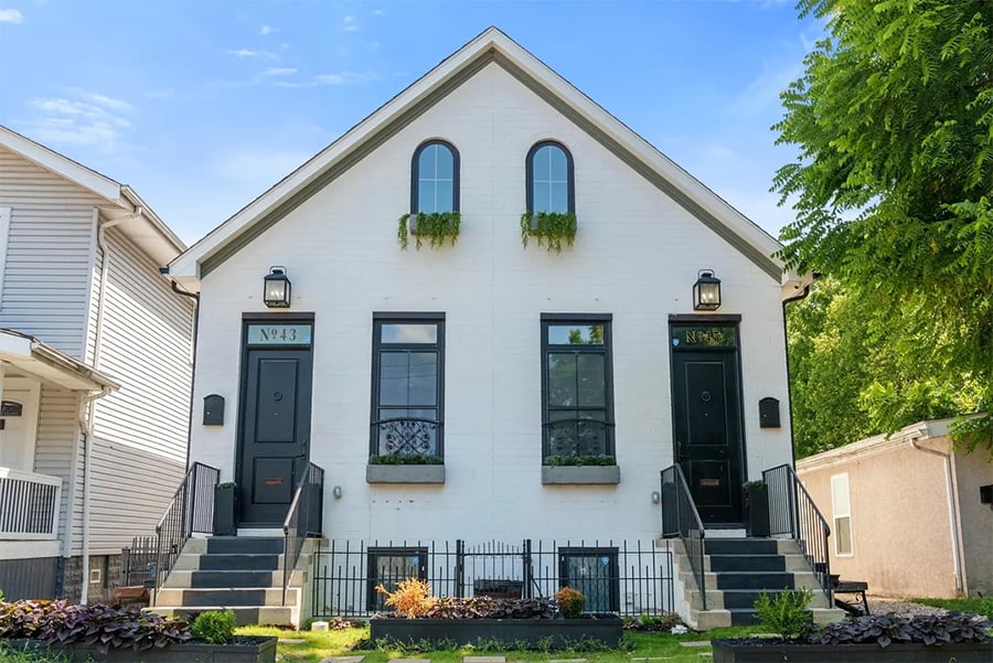 A white duplex house with black trim, two front doors, arched windows above, stairs leading to each entrance, and small gardens in front. Blue sky and green trees are in the background.