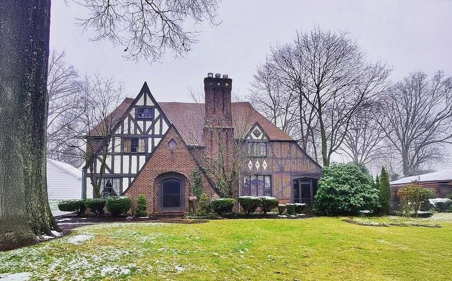A Tudor-style brick house with dark wood trim sits on a lawn scattered with patches of snow, surrounded by leafless trees and bushes under an overcast sky.