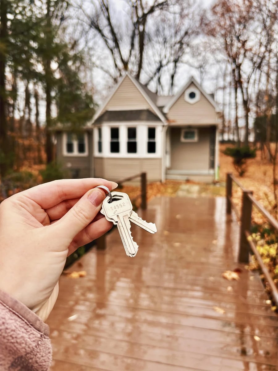 A hand holds keys in front of a small beige house with a porch, surrounded by autumn trees and wet leaves on the ground.