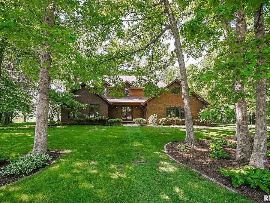 A large brick house with multiple gables sits behind mature trees and green lawn, surrounded by landscaped gardens and shaded by leafy branches on a sunny day.