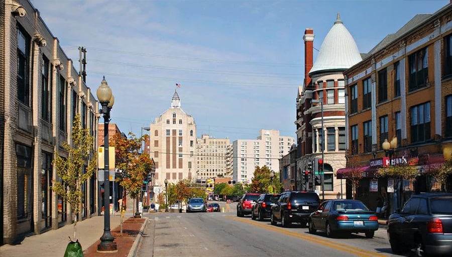 A city street with parked cars, historic and modern buildings, streetlights, trees, and people walking on the sidewalk under a blue sky.