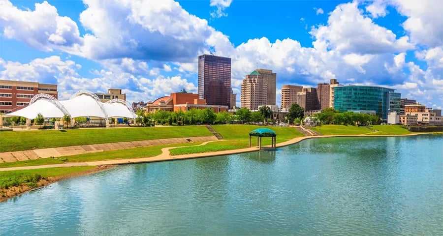 A river curves through a city with green banks, a small dock, white canopies, and tall modern buildings under a blue sky with puffy clouds.