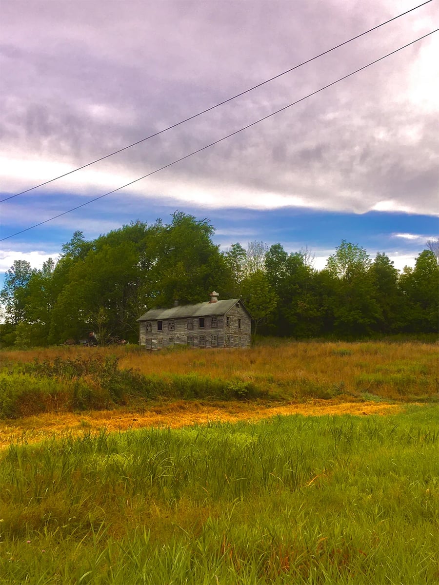 An old, weathered house stands alone in a grassy field, surrounded by trees under a cloudy sky, with power lines crossing above.