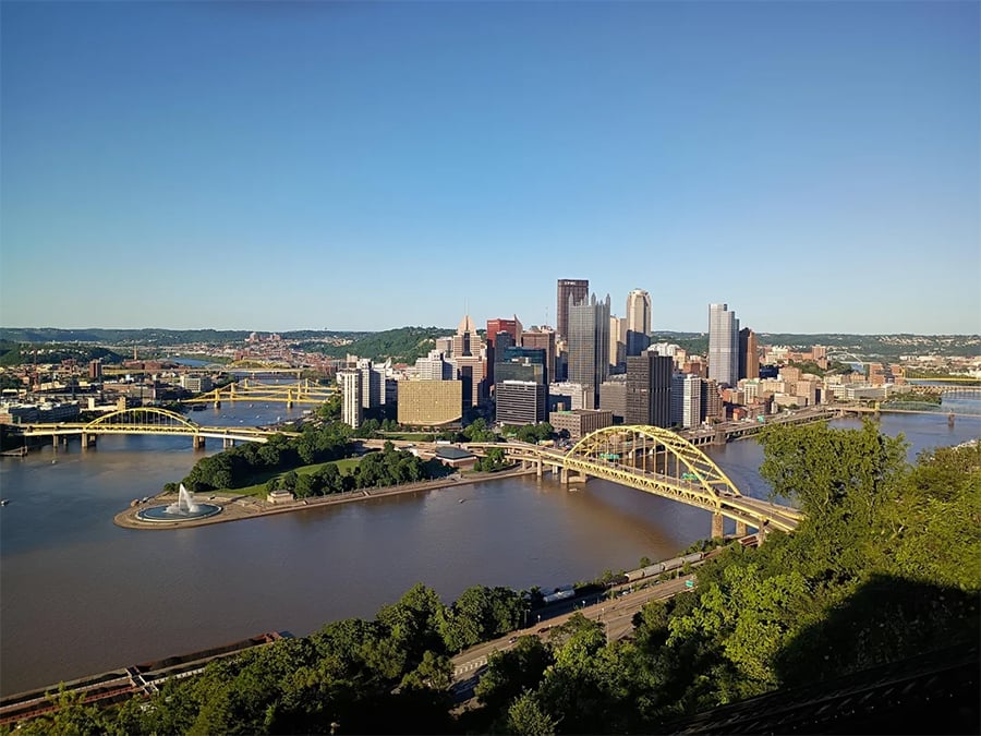 Downtown Pittsburgh skyline with yellow bridges crossing the rivers, a fountain at the river confluence, and clear blue sky in the background.