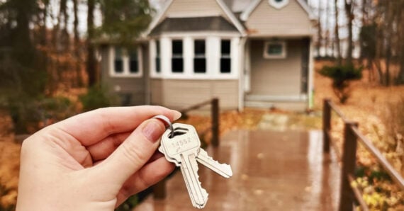 A hand holds two silver keys in focus, with a small beige house and a path leading to it blurred in the background. The scene is surrounded by autumn trees.