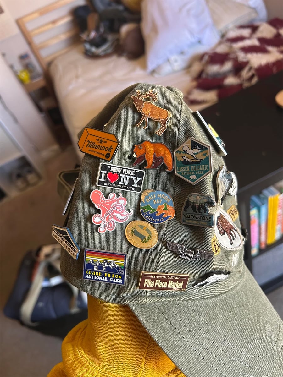 A green baseball cap decorated with various souvenir pins, including national parks, animals, an octopus, “I ❤️ NY,” and Pike Place Market, held indoors with a blurred background of a bed, bookshelf, and duffel bag.