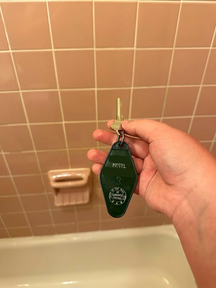 A hand holds a motel room key with a green tag over a pink tiled bathroom wall. Part of a bathtub and a pink soap holder are visible in the background.